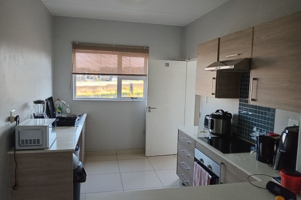 A functional kitchen featuring modern wood-finish cabinetry, integrated appliances including an oven and hob, tiled flooring, and a window with venetian blinds.
