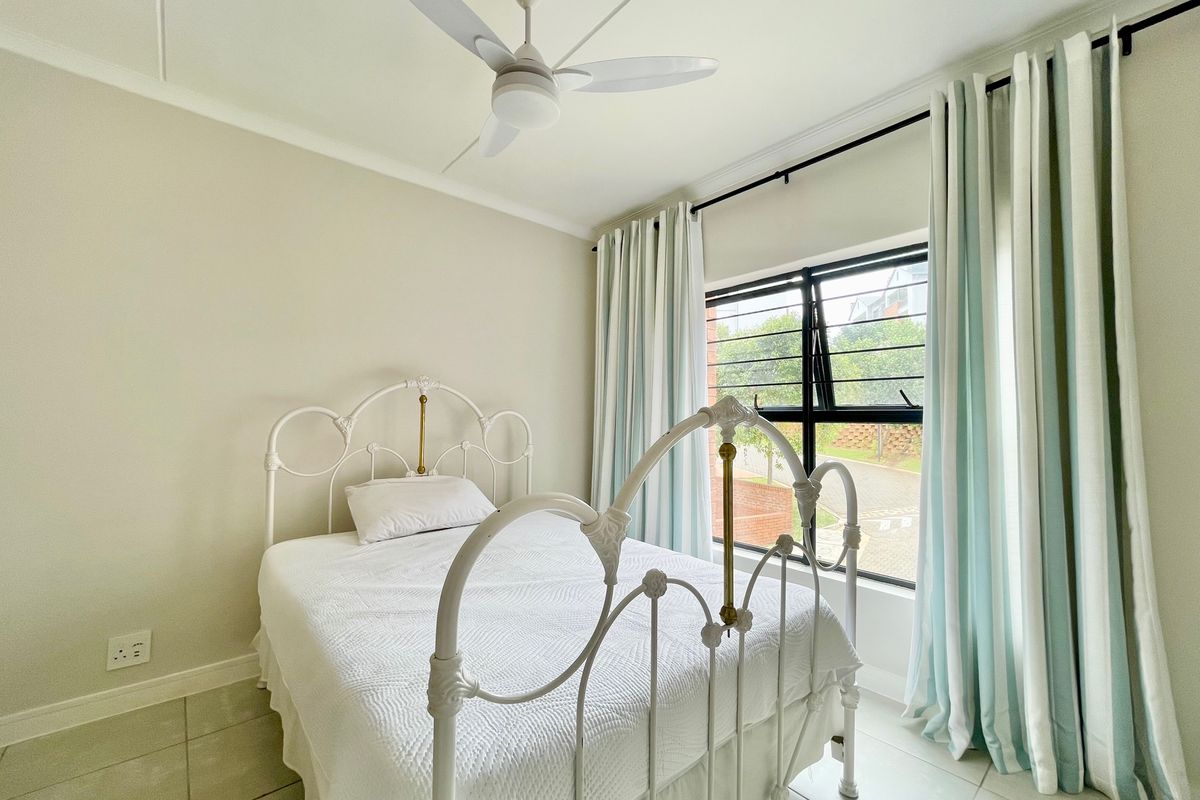 A bright and well-maintained bedroom featuring neutral tiled flooring, a ceiling fan, and large windows with security bars.