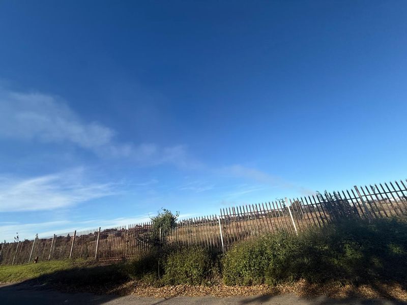 A view of an undeveloped plot of land enclosed by a metal security fence under a clear blue sky.