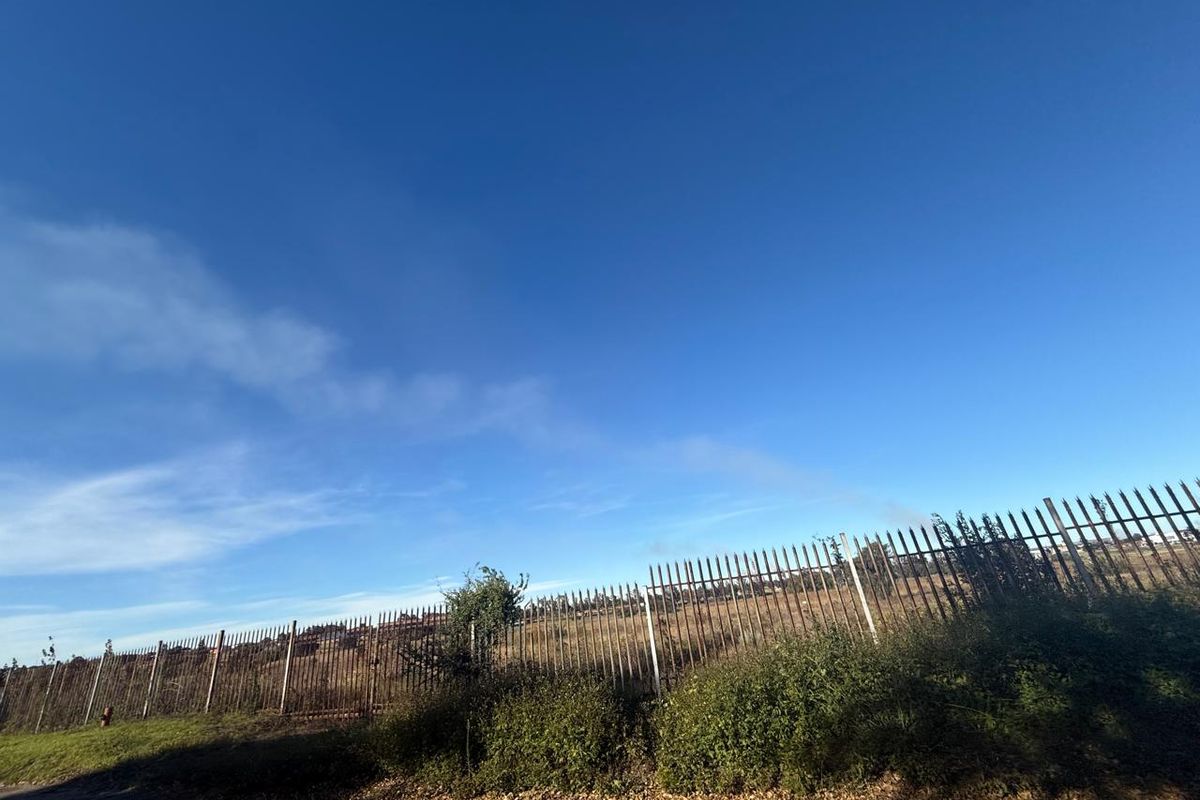A view of an undeveloped plot of land enclosed by a metal security fence under a clear blue sky.
