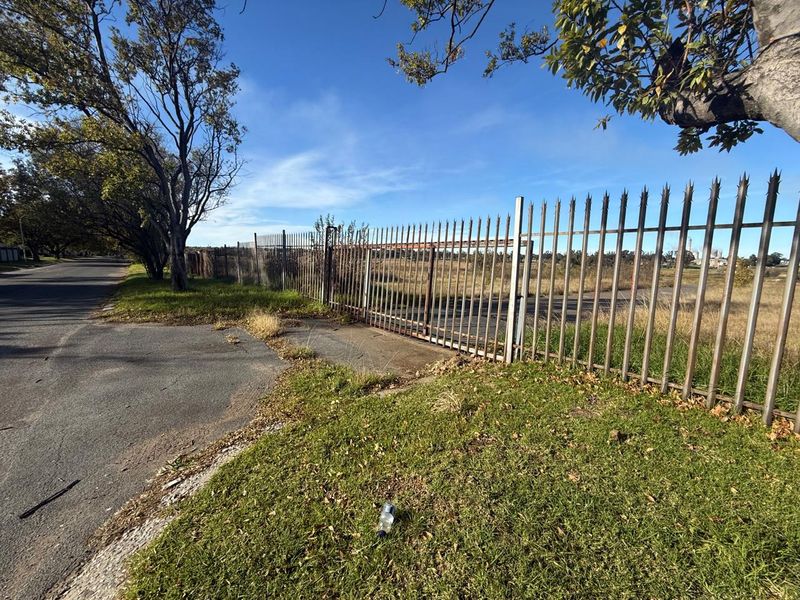 A street-level view of a vacant plot featuring rusted metal security fencing and overgrown grass along the roadside.