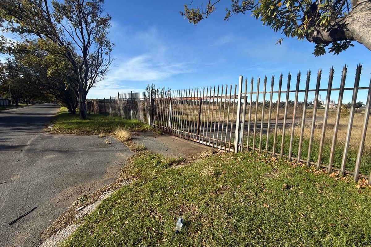 A street-level view of a vacant plot featuring rusted metal security fencing and overgrown grass along the roadside.