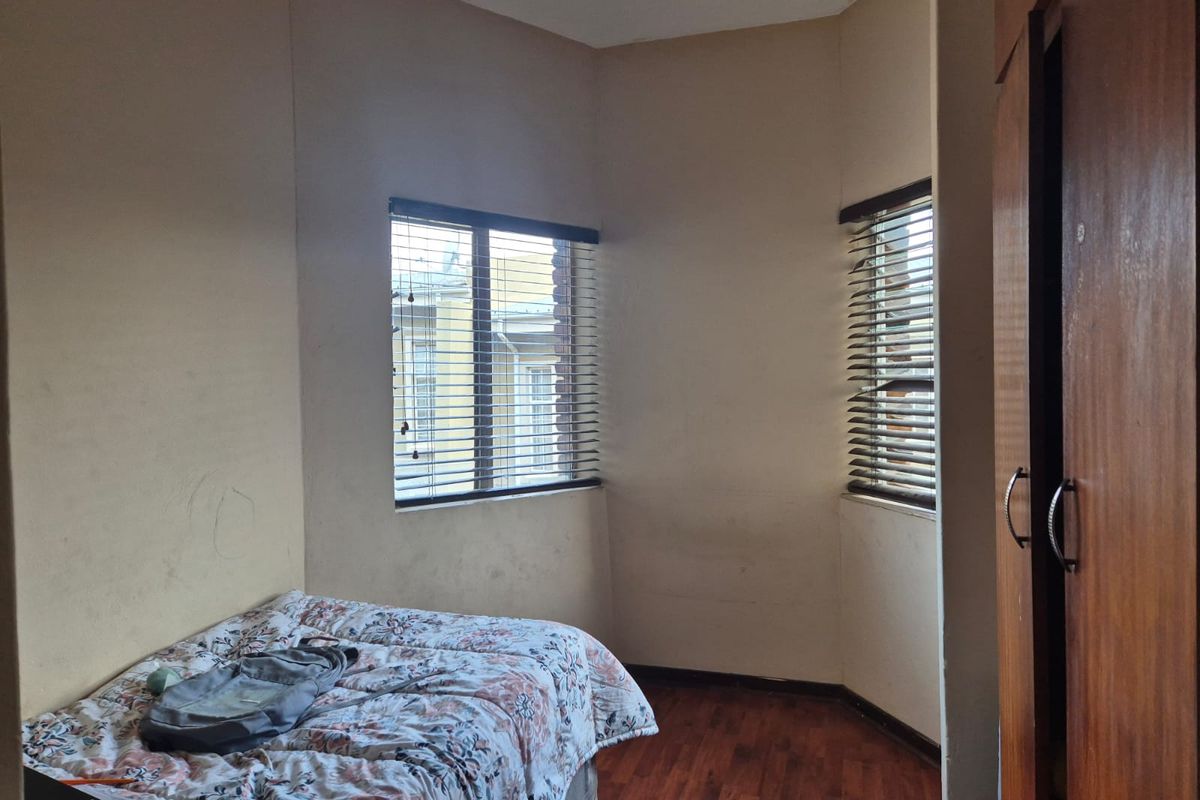 A bedroom featuring laminate flooring, built-in wooden wardrobes, and dual windows with venetian blinds, offering a functional space with potential for cosmetic updates.