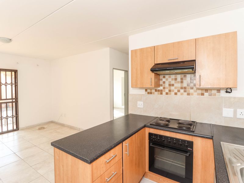 Open-plan kitchen featuring light wood cabinetry, dark countertops, and integrated appliances, adjacent to a tiled living area with security gate access.