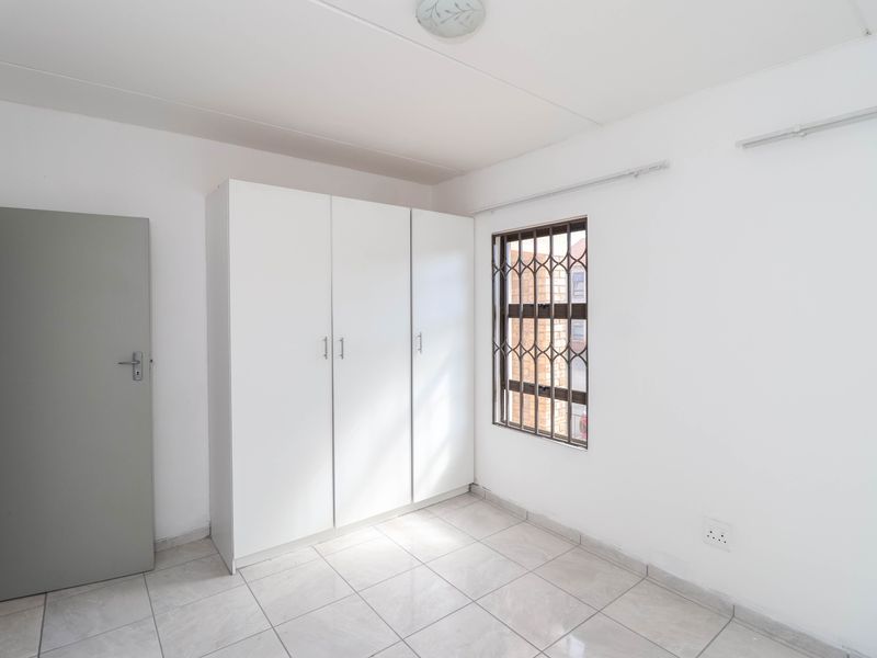 A bright, empty bedroom featuring tiled flooring, a built-in wardrobe, and a window fitted with a security gate.