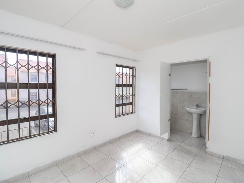 A bright, empty bedroom featuring tiled flooring, security bars on the windows, and an en-suite area with a pedestal basin.