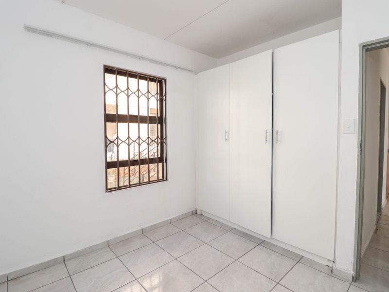 A clean, empty bedroom featuring neutral tiled flooring, built-in cupboards, and a window fitted with a security gate.