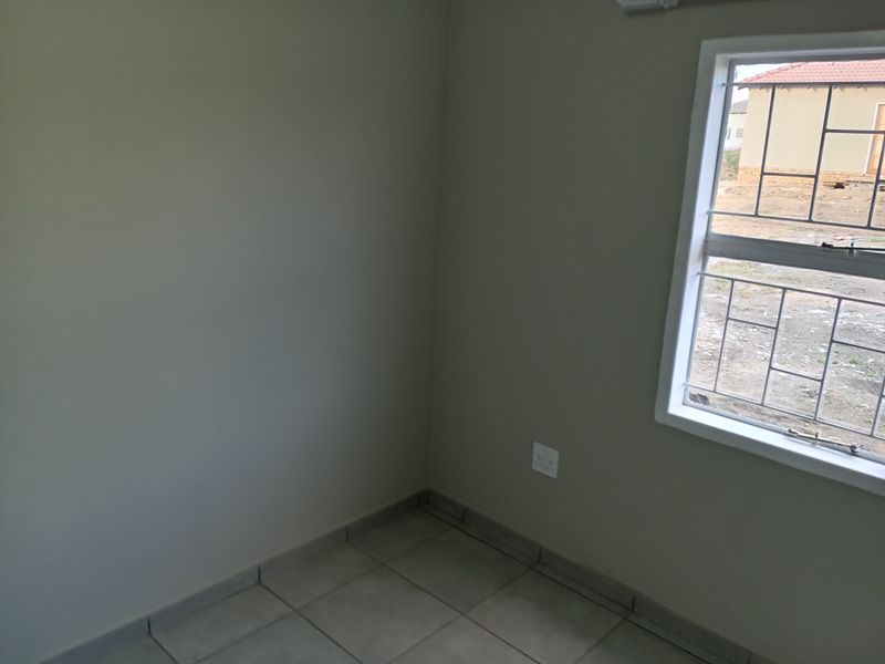 A corner view of an empty bedroom featuring tiled flooring, neutral wall paint, and a window with security bars.