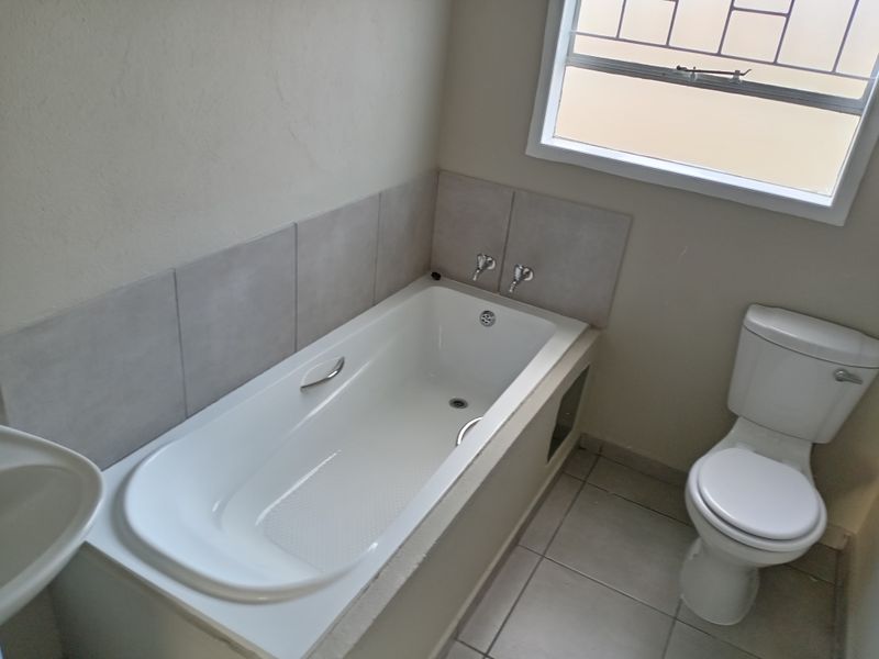 A functional bathroom featuring a built-in bathtub with tiled splashback, a standard toilet, and a window with security bars.