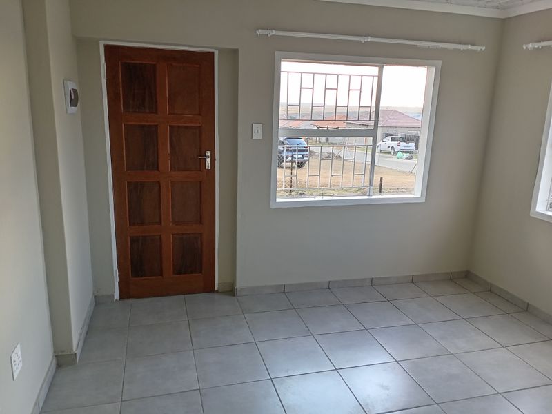 A bright, empty living room featuring neutral ceramic floor tiles, a solid timber door, and large windows fitted with security burglar bars.
