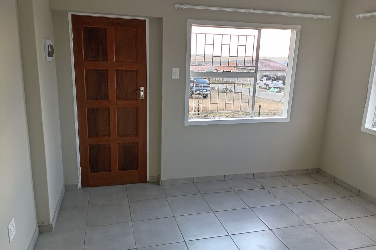 A bright, empty living room featuring neutral ceramic floor tiles, a solid timber door, and large windows fitted with security burglar bars.