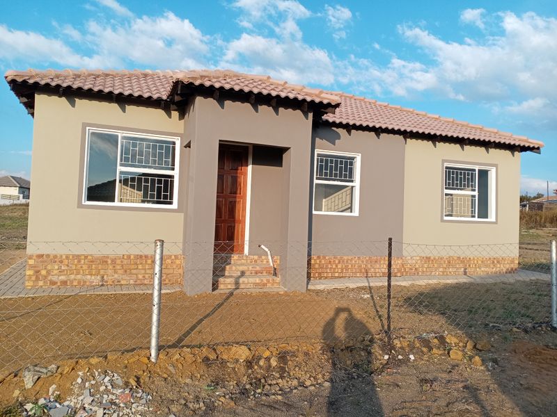 A modern single-story house featuring a tiled roof, brick base detailing, and secure window bars, set on an undeveloped plot.