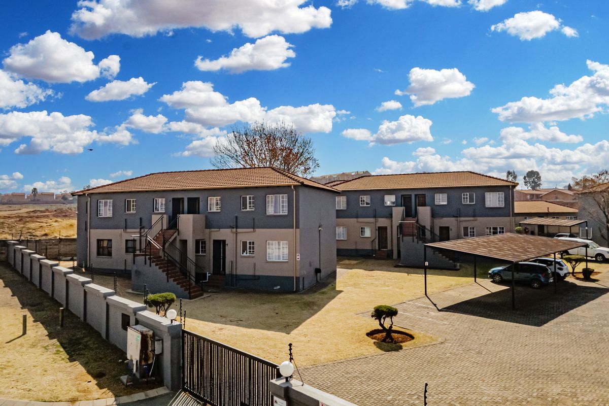 Aerial view of a multi-unit apartment complex featuring paved parking, carports, and perimeter security walls under a clear blue sky.