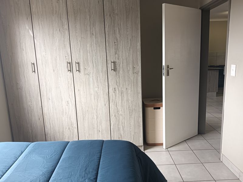 A bedroom featuring light wood-grain built-in cupboards and neutral tiled flooring, with a doorway leading to an adjacent living area.
