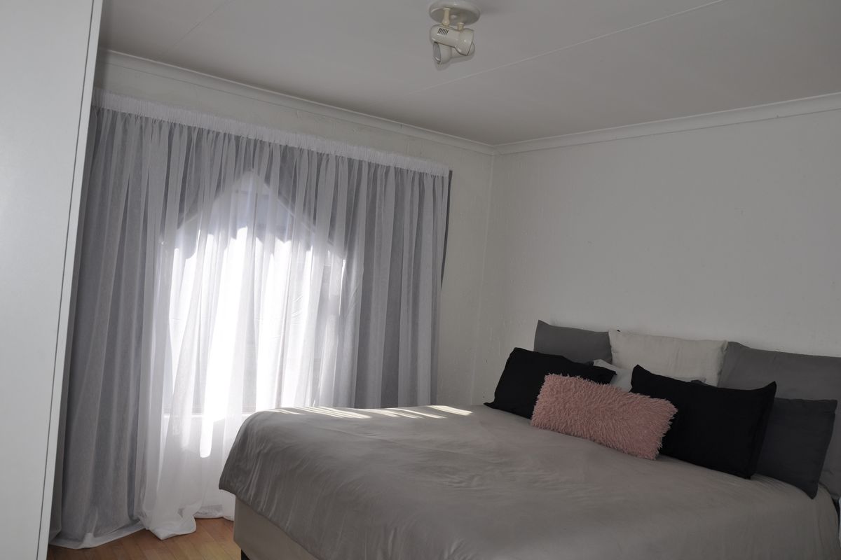 A bright bedroom featuring neutral walls, wooden flooring, and a large window dressed with sheer curtains, providing a clean and simple living space.