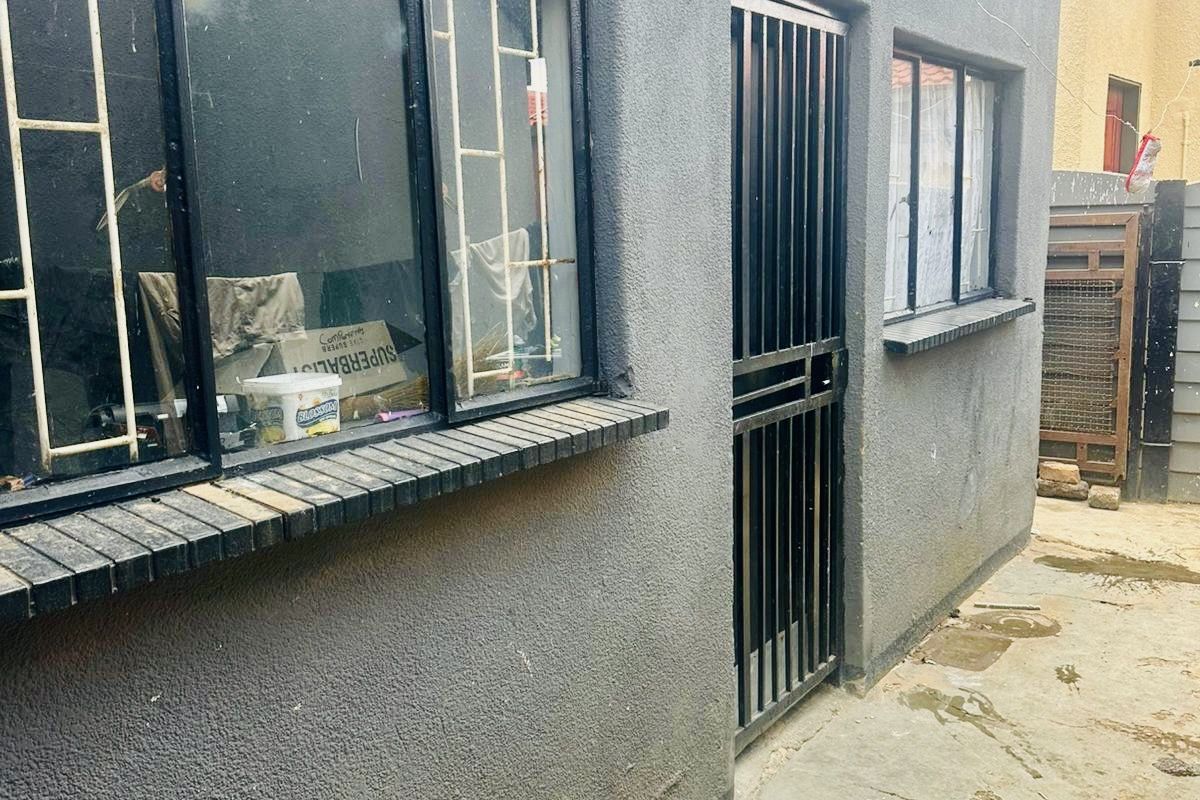 Rear exterior view of the property featuring a security gate, burglar bars, and concrete paving with visible signs of wear and maintenance requirements.