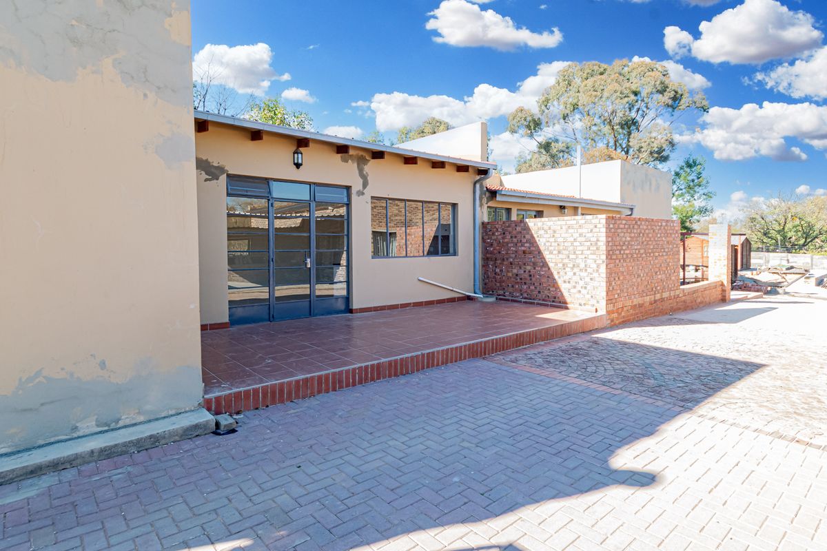 Rear exterior view featuring a tiled patio, brick walling, and paved driveway area with visible maintenance required on the exterior plaster and paintwork.