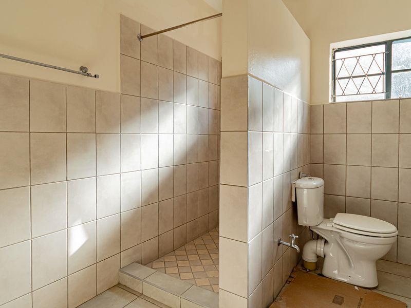 A functional bathroom featuring a walk-in shower, toilet, and pedestal basin, finished with neutral ceramic tiling and security-barred windows.
