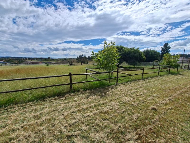 A view of a vacant residential plot featuring open grassland and timber post and rail fencing under a cloudy sky.