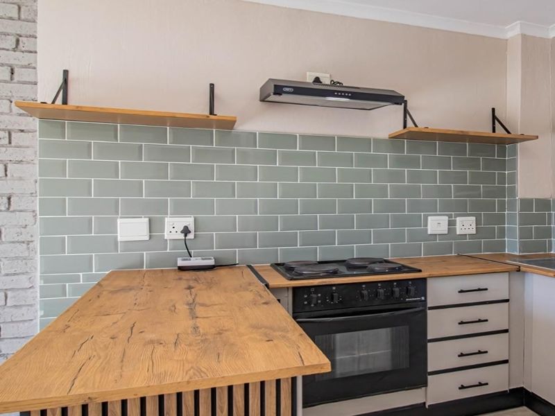 Modern kitchen featuring a stylish sage green subway tile backsplash, warm wooden countertops, built-in oven with hob, and exposed brick wall detailing.