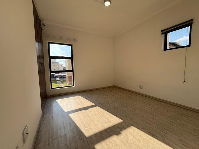 A bright, empty bedroom featuring modern laminate flooring, built-in cupboards, and aluminium-framed windows, offering a clean and neutral space ready for personalization.