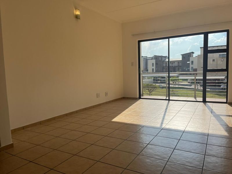 An empty, sunlit living room featuring neutral tiled flooring and a large sliding glass door that opens onto a balcony with neighborhood views.