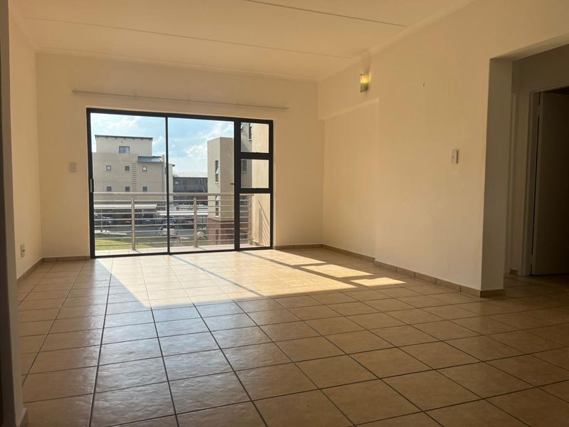 A bright, empty living room featuring neutral tiled flooring and a large sliding glass door that opens onto a balcony.