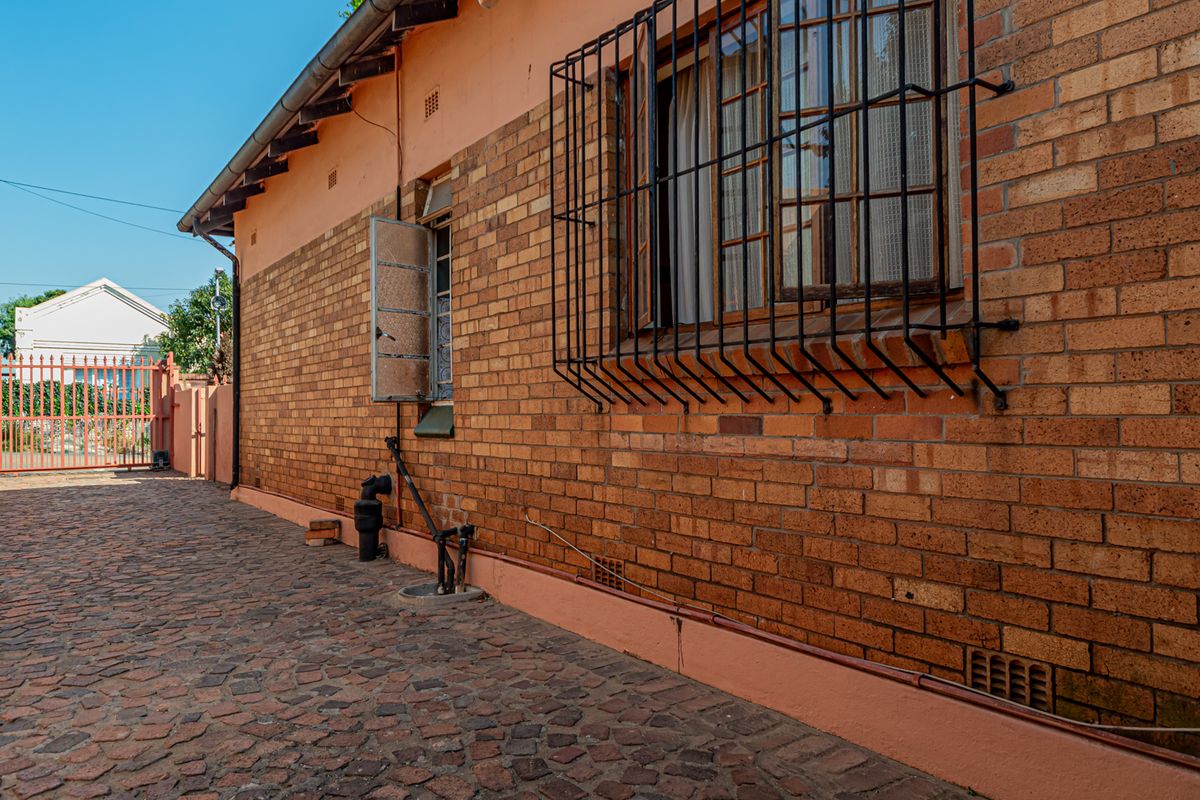 Side exterior view of the house featuring face brick walls, security bars on windows, and cobblestone paving leading to a metal gate.