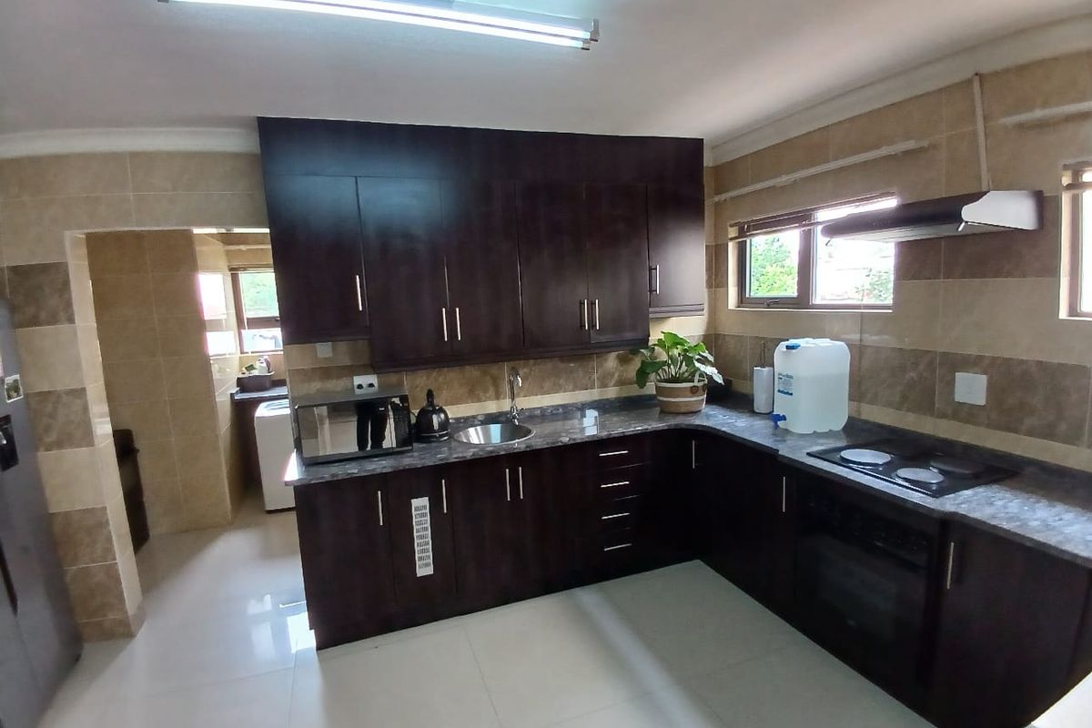 A functional kitchen featuring dark wood cabinetry, granite countertops, built-in oven, electric hob, and tiled splashbacks, with an adjacent scullery area.