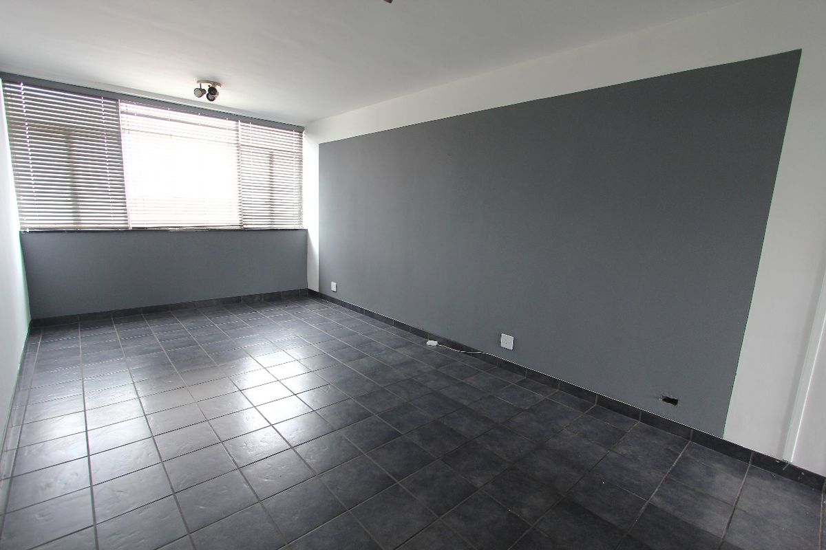An empty living room featuring dark tiled flooring, a large window with venetian blinds, and a grey accent wall.