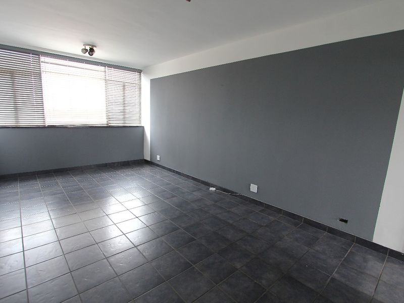 An empty living room featuring dark tiled flooring, a large window with venetian blinds, and a grey accent wall.