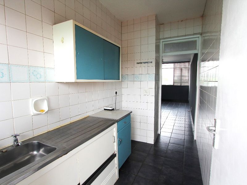 A compact, dated kitchen featuring built-in cabinetry, a stainless steel sink, and tiled finishes, leading into an adjacent living area.