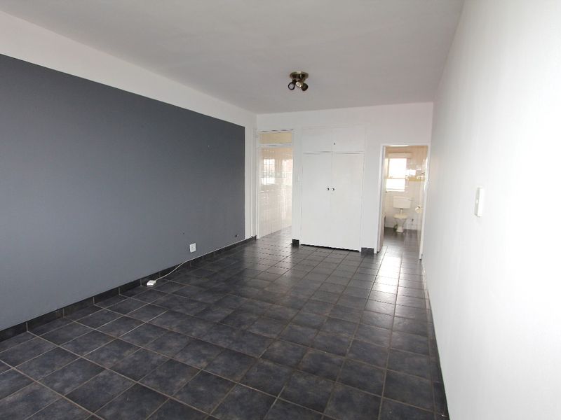 An empty living room featuring dark tiled flooring, a grey accent wall, and built-in storage cupboards, with a view into the adjacent bathroom.
