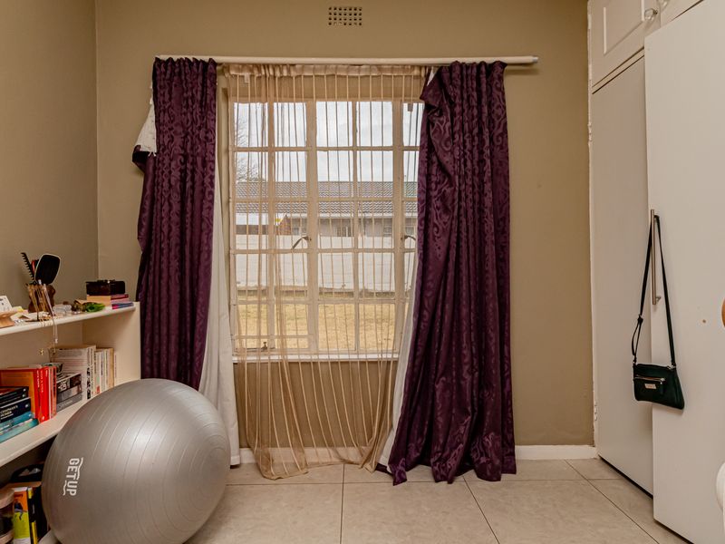 A bedroom featuring tiled flooring, built-in cupboards, and a window with security bars, offering a functional space with neutral wall tones.