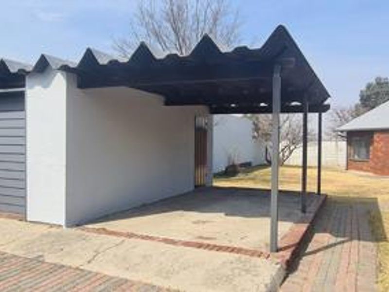 An open-sided carport structure featuring a corrugated roof, concrete flooring, and brick paving, providing covered parking space.