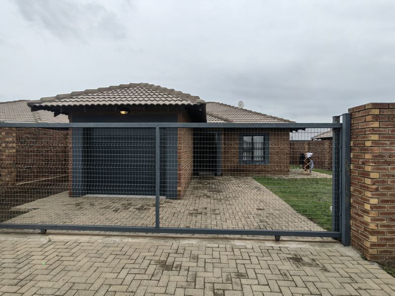 Front view of a single-story brick house featuring a paved driveway, integrated garage, and secure perimeter fencing.