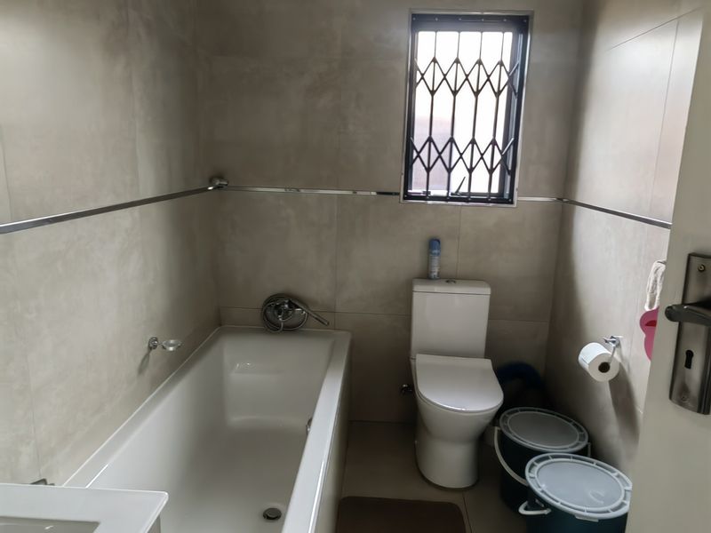 A functional bathroom featuring neutral wall and floor tiles, a built-in bathtub, a standard toilet, and a vanity basin with a security-grilled window.