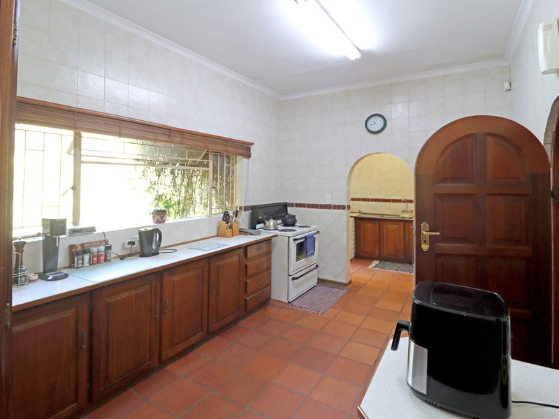 A functional kitchen featuring wooden cabinetry, terracotta floor tiles, and a tiled splashback, with an adjoining scullery area accessible through an arched doorway.
