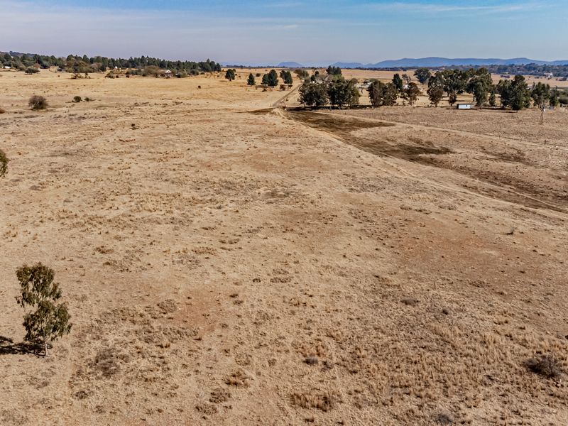 An aerial view of a large, expansive vacant residential plot featuring dry grassland, scattered trees, and distant mountain views.
