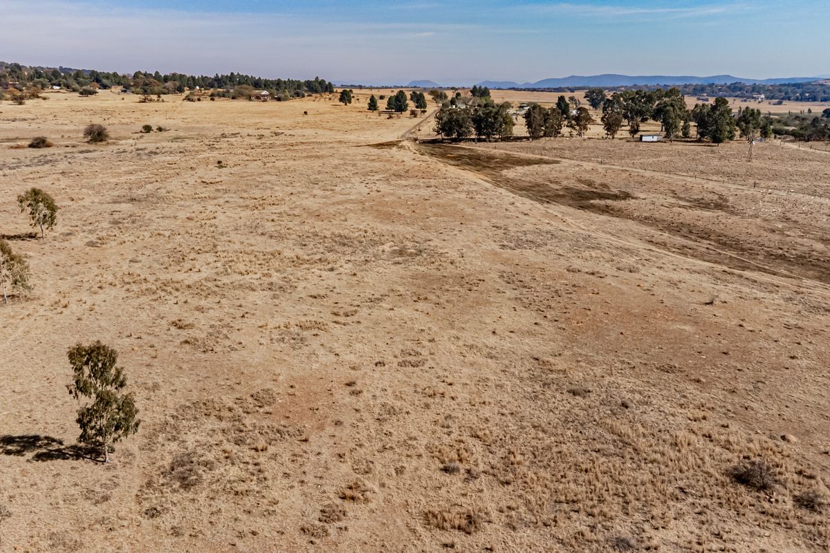 An aerial view of a large, expansive vacant residential plot featuring dry grassland, scattered trees, and distant mountain views.