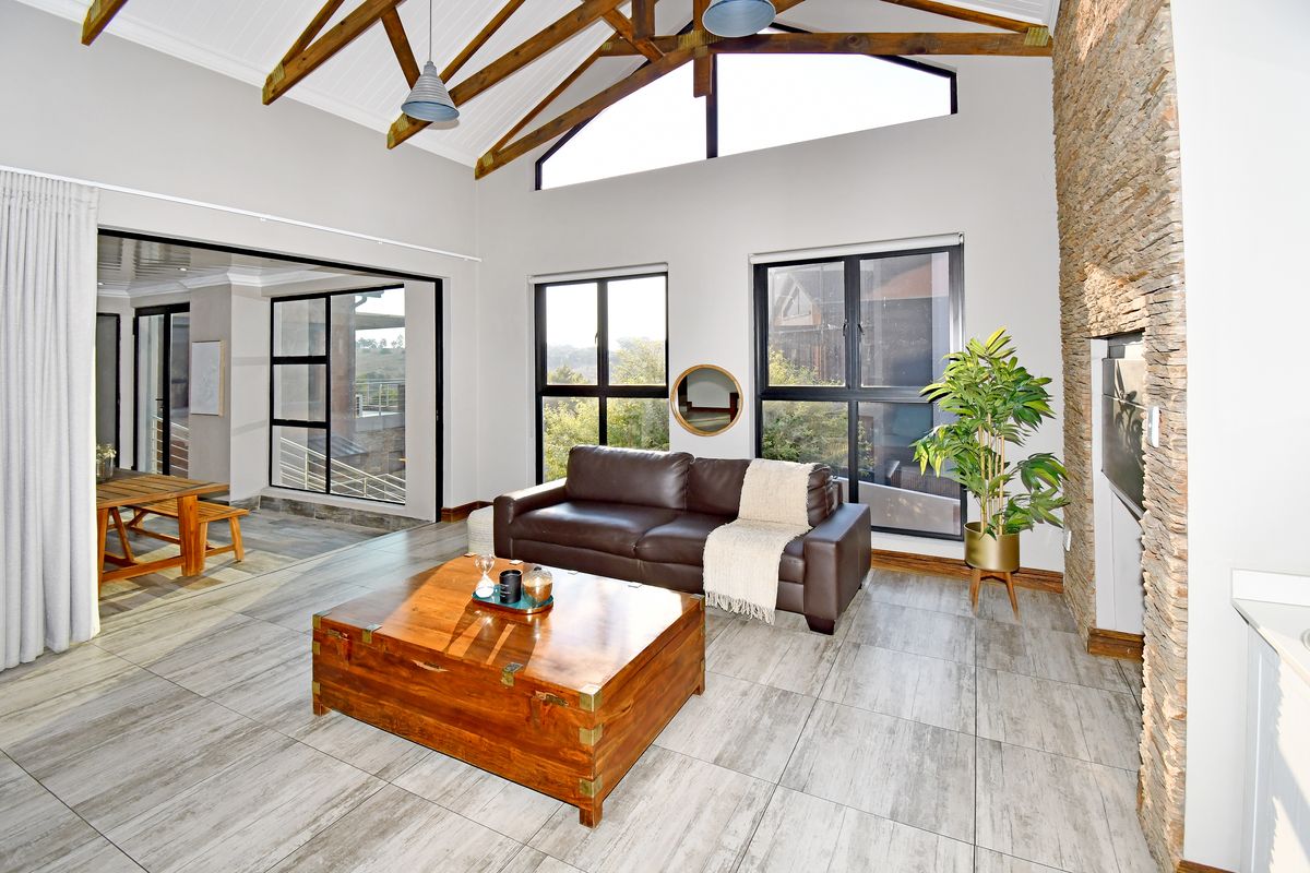 Spacious living room featuring vaulted ceilings with exposed timber trusses, stone-clad fireplace, large windows, and modern tiled flooring.