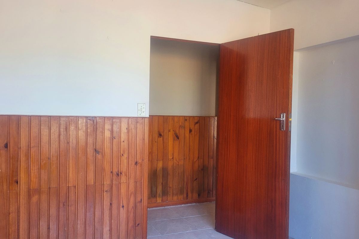An empty bedroom featuring dated timber wall paneling, a solid wood door, and tiled flooring, offering a blank canvas for renovation.