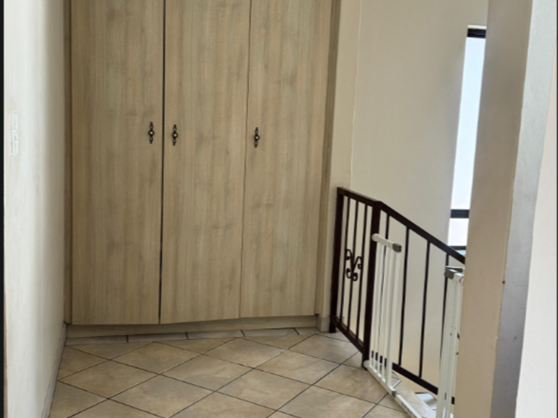 A bright hallway featuring neutral tiled flooring, built-in wooden cupboards for storage, and a metal balustrade overlooking the staircase.
