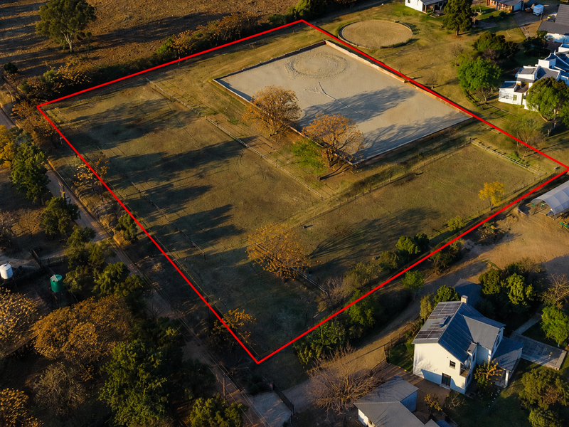 Aerial view of a large residential plot featuring a fenced perimeter, a sand riding arena, a round pen, and established trees.