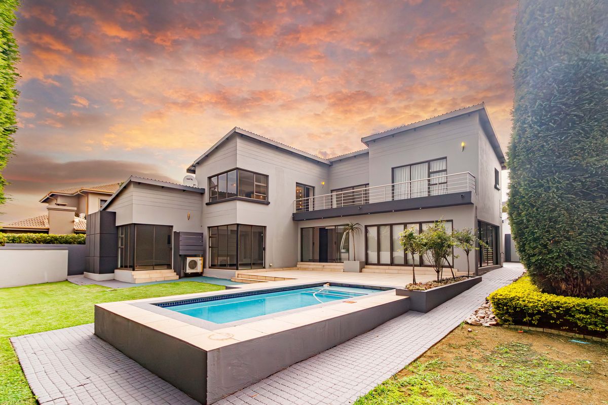 Rear view of a modern two-story house featuring a swimming pool, paved patio area, and an upper-level balcony with glass railings.