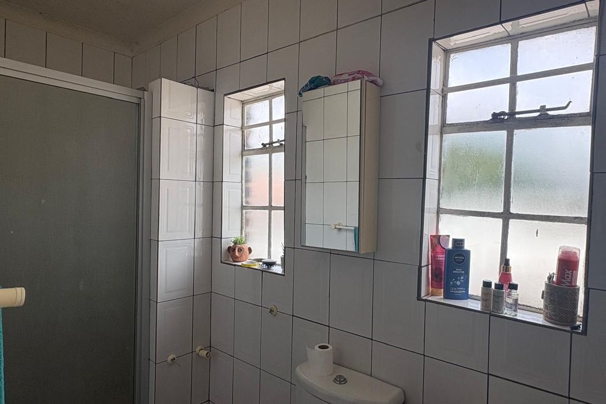 A dated bathroom featuring white wall tiles, a shower enclosure, a toilet, and two windows with frosted glass panes.