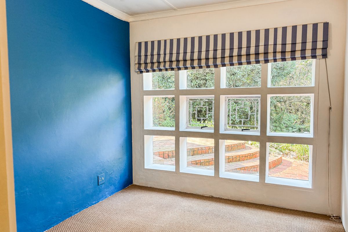 An empty bedroom featuring a distinctive grid-pattern window overlooking a garden, carpeted flooring, and a bold blue accent wall.