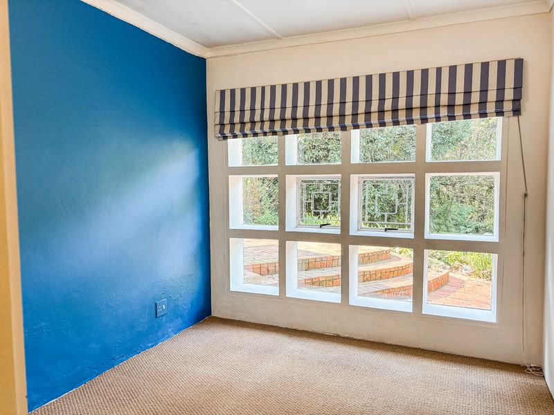 An empty bedroom featuring a distinctive grid-pattern window overlooking a garden, carpeted flooring, and a bold blue accent wall.