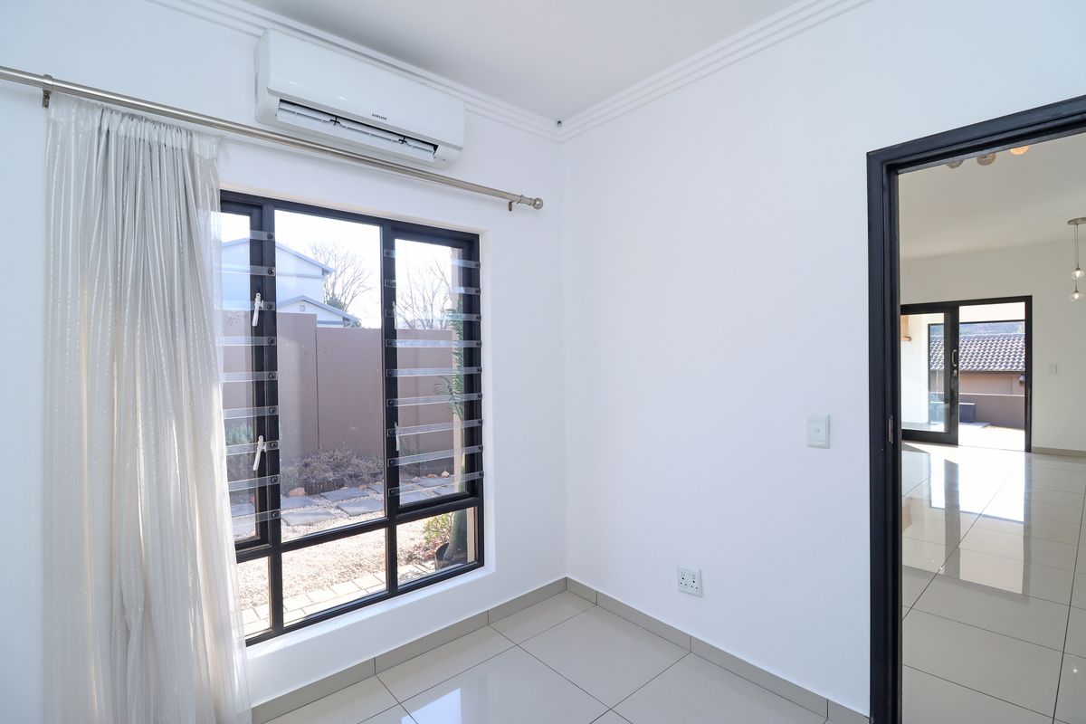 A bright, empty bedroom featuring tiled flooring, modern louvre windows, and a wall-mounted air conditioning unit, with a view into the adjacent living area.