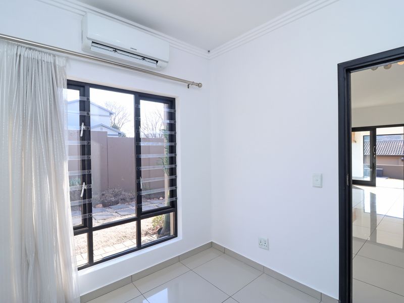 A bright, empty bedroom featuring tiled flooring, modern louvre windows, and a wall-mounted air conditioning unit, with a view into the adjacent living area.